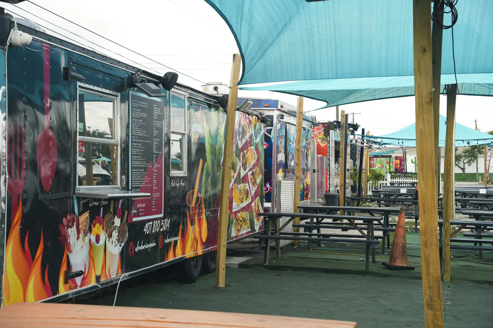 View of an outdoor dining area with food trucks. There are picnic tables and seating for customers to enjoy their meals. The atmosphere is casual, social, and lively.