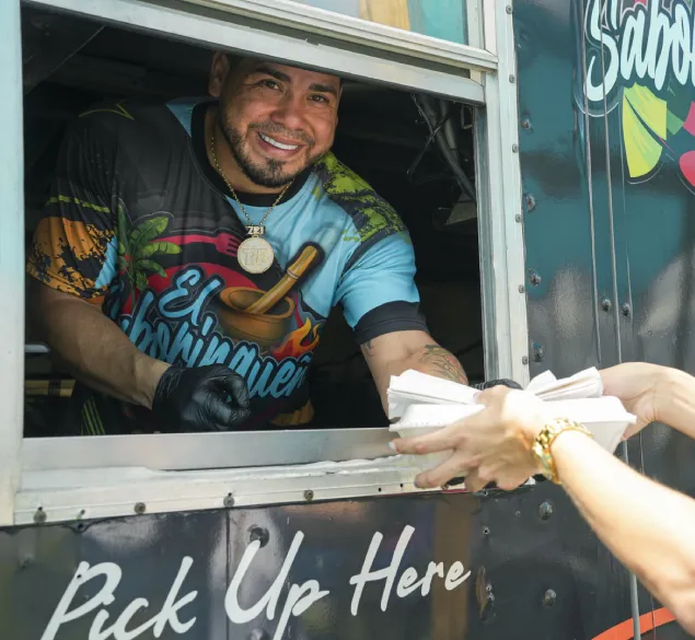 A friendly, smiling worker at a food truck hands a plate of food to a customer. The interaction appears cordial and cheerful, highlighting the positive experience of shopping at a food truck.