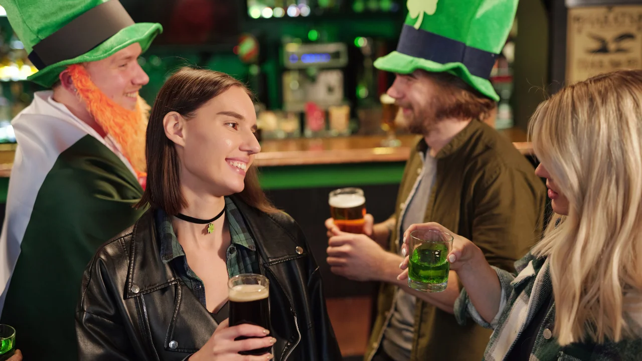 Four joyful friends celebrating in a bar. They're wearing leprechaun hats and holding drinks, conveying a festive, St. Patrick's Day atmosphere.
